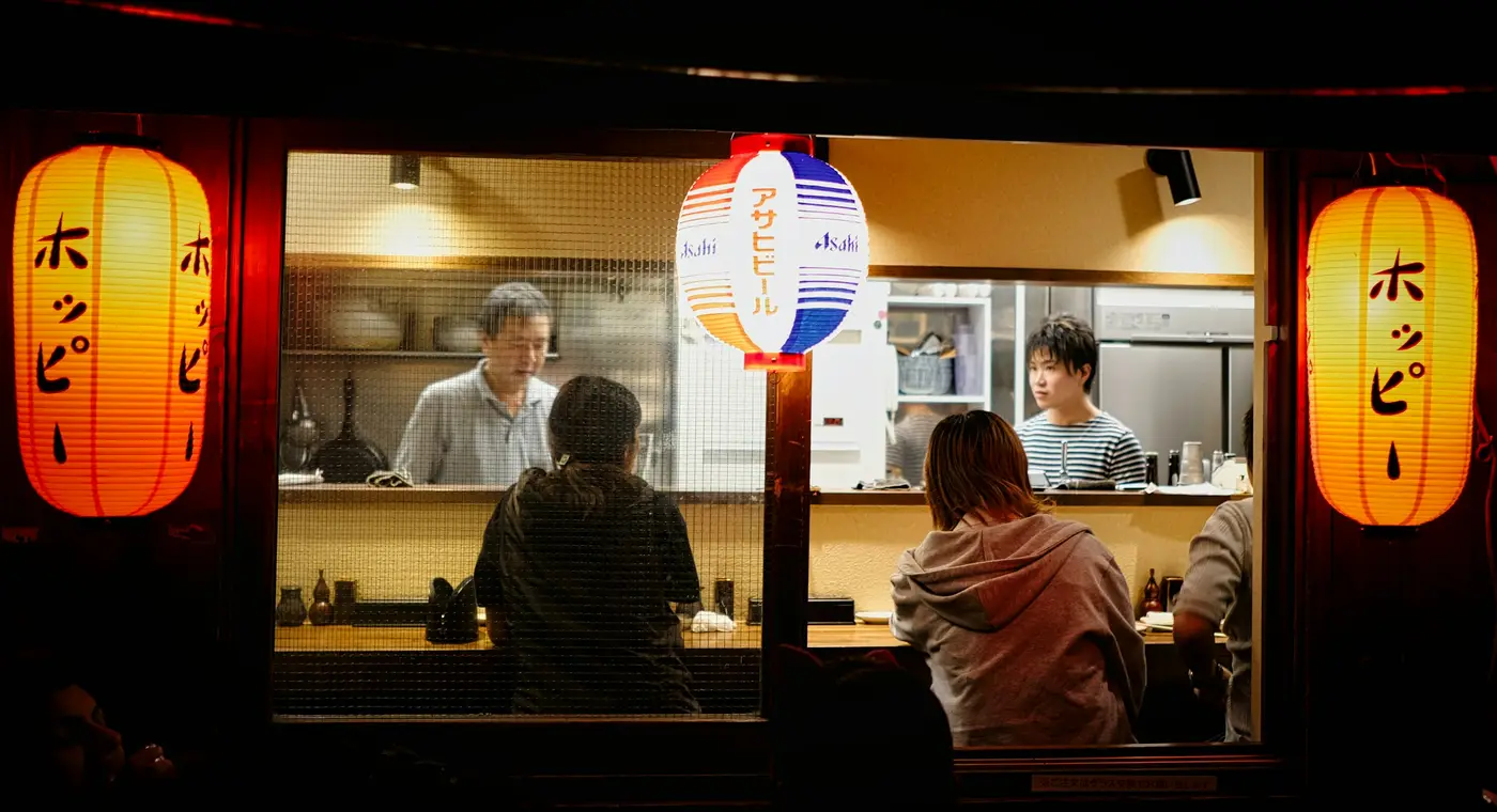 Diners at the counter of a small izakaya, seen through the window from the street at night