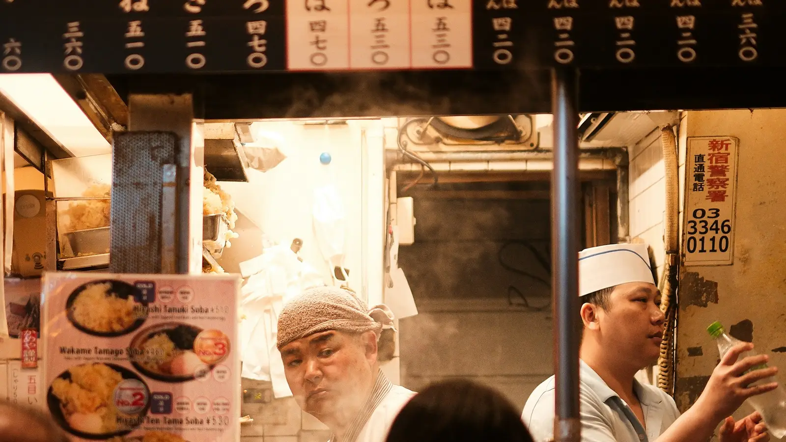 Cooks at a tiny noodle counter in Shinjuku, with the shop's phone number on the wall